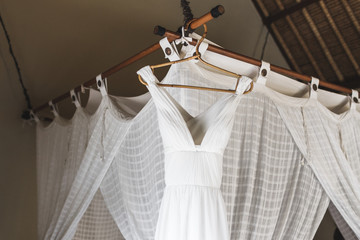 White wedding dress hanging inside room on baldachin with curtains. Bali style