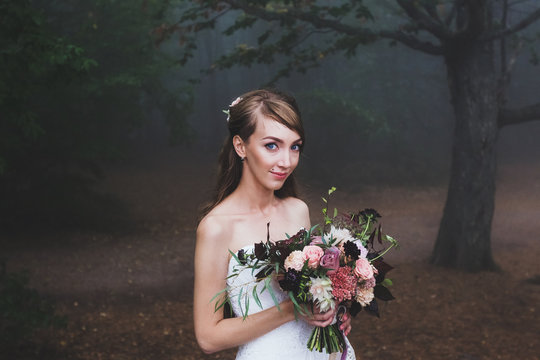 Stylish young bride with piercings holding wedding bouquet in pink and dark tones. Forest around