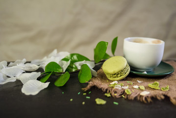 White cup of coffee on a green plate with macaroons and flower petals and wood background with linen