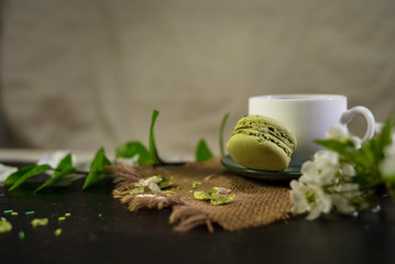 White cup of coffee on a green plate with macaroons and flower petals and wood background with linen