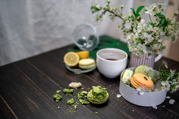 Crushed pastel, orange and green macaroons on a wood background with whole one in white plate with jug with flowers on the back