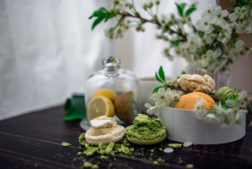 Crushed pastel, orange and green macaroons on a wood background with whole one in white plate with jug with flowers on the back