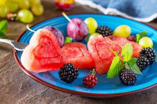 Various fresh fruits and berries on a plate on a brown stone or slate background - watermelon, blackberries, plums and grapes.