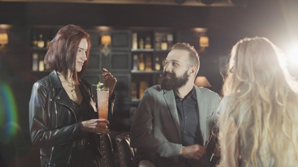 Company of friends talking in night bar. Redhead woman is giving high five to her bearded friend in bar, then they clink glasses together.
