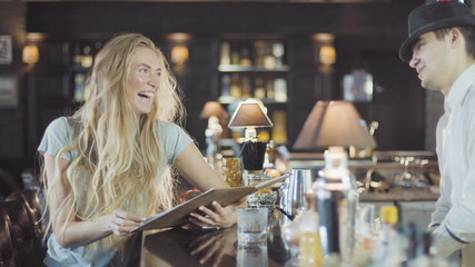 Bartender helps to blonde 20s woman make a choice in menu of bar