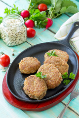 Domestic meat cutlet with white beans and fresh herbs. Cast-iron frying pan full of delicious fried meat cutlets, fresh greens and vegetables on the kitchen table.