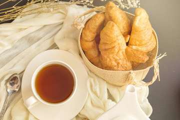 Freshly baked croissants In the basket and cup of tea on a dark table background. in a windows light. Breakfast concept