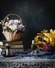 assorted flowers in a basket and on old table with books, perfume bottle, candle and textile on a green background with vignette, toned for a vintage effect