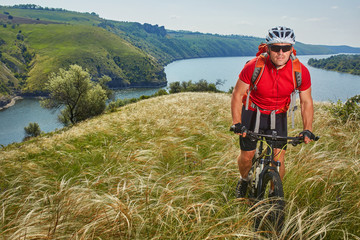 Attractive cyclist riding mountainbike on the meadow above river in summer season in the countryside.