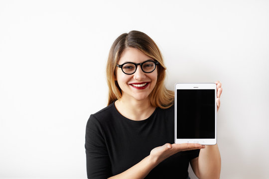 Charming young businesswoman with pretty smile wearing black dress and eyeglasses looking at camera and showing a digital tablet computer with blank copy screen for your advertising content.