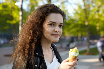 Headshot of pretty girl standing outdoors holding waffle cone, eating delicious fruit ice-cream during walk in city park, looking at camera with cute charming smile. People and lifestyle concept