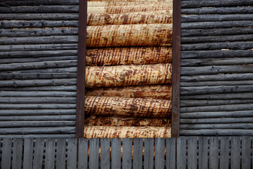 Old weathered log barn wall facade fragment and batten fence of rustic horizontal and vertical grey timber background loaded with freshly cut debarked pine wood lumber.