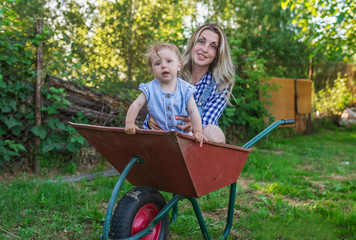 Mother and her whimsical little daughter sit in a wheelbarrow outdoors