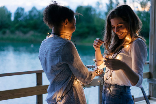 Two women standing face to face, illuminated fairy lights wrapped around their neck, smiling