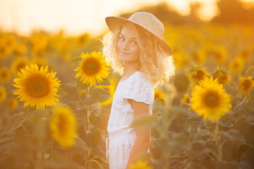 Pretty curly girl in white dress in filed of sunflowers over sunset lights