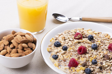 Cereal bowl breakfast with milk, raspberries and blueberries next to orange juice and bowl with almonds.