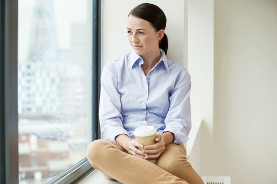 Businesswoman sitting on window ledge, looking out of window, holding takeaway coffee cup