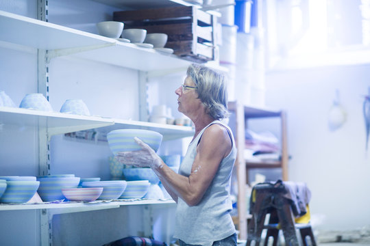 Senior woman in pottery workshop, placing bowl on shelf