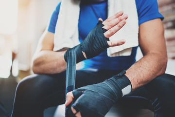 Obraz premium Muscular Boxer man prepairing hands for hard kickboxing training session in gym. Young athlete tying black boxing bandages.Blurred background. Horizontal.