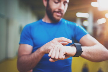 Young Muscular bearded athlete checking burned calories on electronic smart watch application after good indoor workout session in fitness gym.Blurred background.