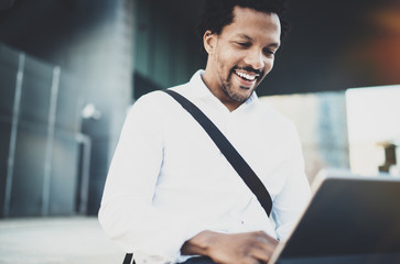 Fototapeta premium Smiling young African American man sending email via electronic touch pad pro holding in hands.Concept of guy using Internet-enabled electronic device outside.Blurred background.Cropped.