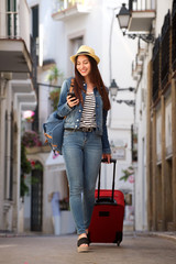 Full length woman walking on street with luggage holding mobile phone