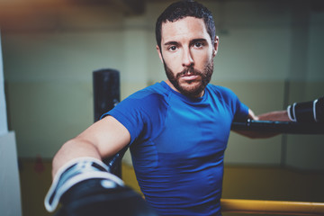 Young boxing man ready to muay thai fight on ring.Bearded athlete in black gloves looking at the camera.Blurred background.Horizontal.