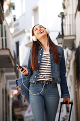 Happy woman walking with earphones and luggage outside
