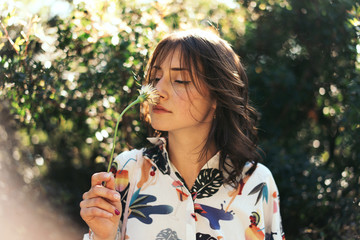 Charming caucasian female with brown hair is smelling the flower while standing on a blurred natural background. Young woman in colorful t-shirt is looking at flower while relaxing at the city park - 163778004