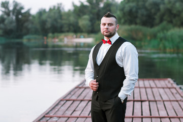 Young stylish man in a waistcoat, horizontal portrait of the groom, portrait on a background of nature, the river and the pier, look at the camera