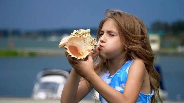 The girl costs with a cockleshell in hands at a pier with boats. the girl plays with a big cockleshell. The girl blows in a big cockleshell as in a horn