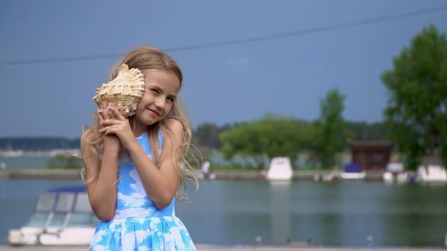A bit the fair-haired girl listens to a surf of waves in a big cockleshell. The girl costs with a cockleshell in hands at a pier with boats