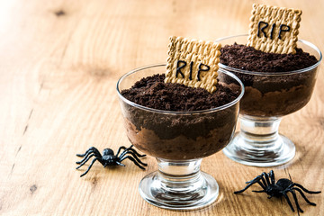 Funny Halloween chocolate mousse with tomb cookie and spiders on wooden table

