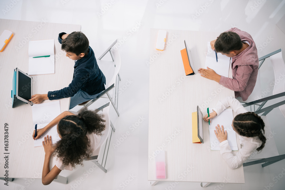 Wall mural overhead view of pupils using digital tablets while sitting at desks in classroom