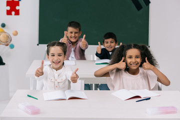 Cheerful multiethnic schoolkids showing thumbs up while sitting at desks in classroom