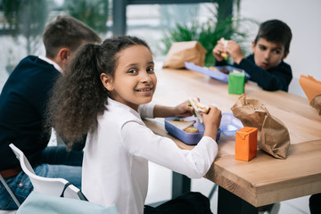 Cute multiethnic schoolkids eating lunch while sitting at table