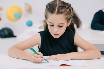 Close-up portrait of cute concentrated schoolgirl writing in exercise book at lesson