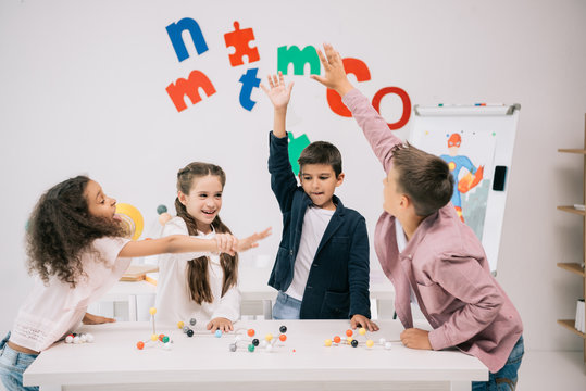 Multiethnic schoolkids giving high five while studying with molecular model in classroom
