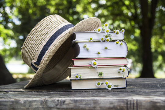 A stack of books on an old wooden table next to a straw hat of a canoe and a bouquet of wild flowers