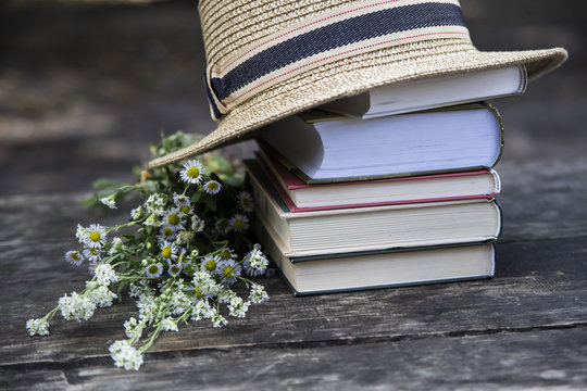 A stack of books on an old wooden table next to a straw hat of a canoe and a bouquet of wild flowers
