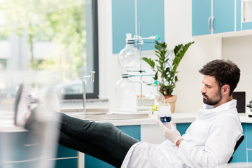 Side view of bearded male scientist holding flask with reagent while sitting in chemical lab