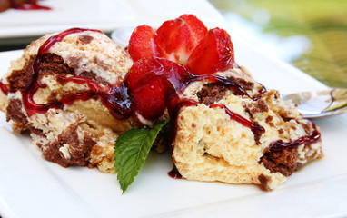 Slice of creamy biscuit cake with cocoa, jam and fresh strawberries and a mint leaf on a white plate.