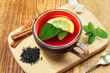 Hot drink in a rustic clay cup made of black tea, fresh mint leaves and lemon on a wooden table. Delicious healthy beverage made of herbals and fruits.