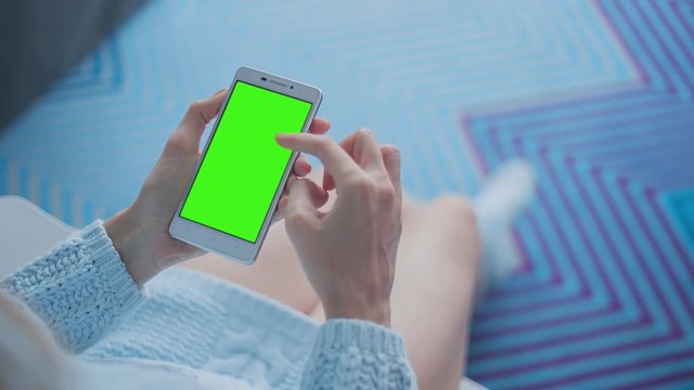 Young Woman in blue sweater sitting on white chair uses SmartPhone with pre-keyed green screen. Few types of gestures - scrolling up and down, tapping, zoom in and out. Perfect for screen compositing