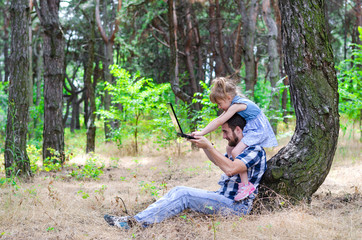 Naklejka na ściany i meble Father and daughter are sitting on nature with a laptop, a bright summer photo with free space.