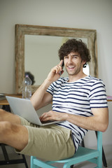 Portrait of a man with mobile phone and laptop. Shot of a happy young man using his laptop and talking with somebody while relaxing at home. 