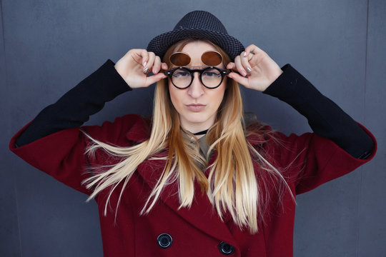Stylish blonde with beautiful makeup in a red coat, hat and black glasses on the background of a gray wall