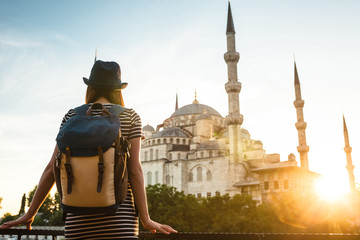Young beautiful girl traveler in a hat with a backpack looking at a blue mosque - a famous tourist...