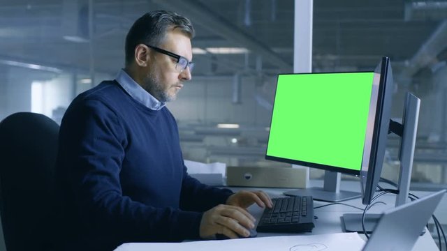 Chief Male Engineer Working on Technical Project on His Person Computer. Second Display Shows Mock-up Green Screen. Out of the Office Window Big Factory is Seen. 4K UHD.