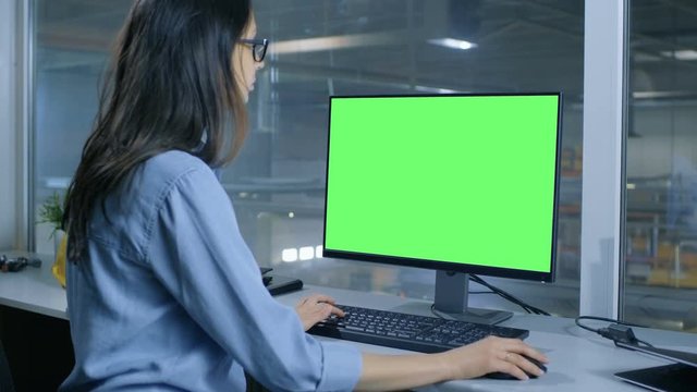  Female Industrial Engineer Works on Her Computer with  Mock-up Green Screen. Inside of the Factory is Seen From Her Office Window.Shot on RED EPIC-W 8K Helium Cinema Camera.